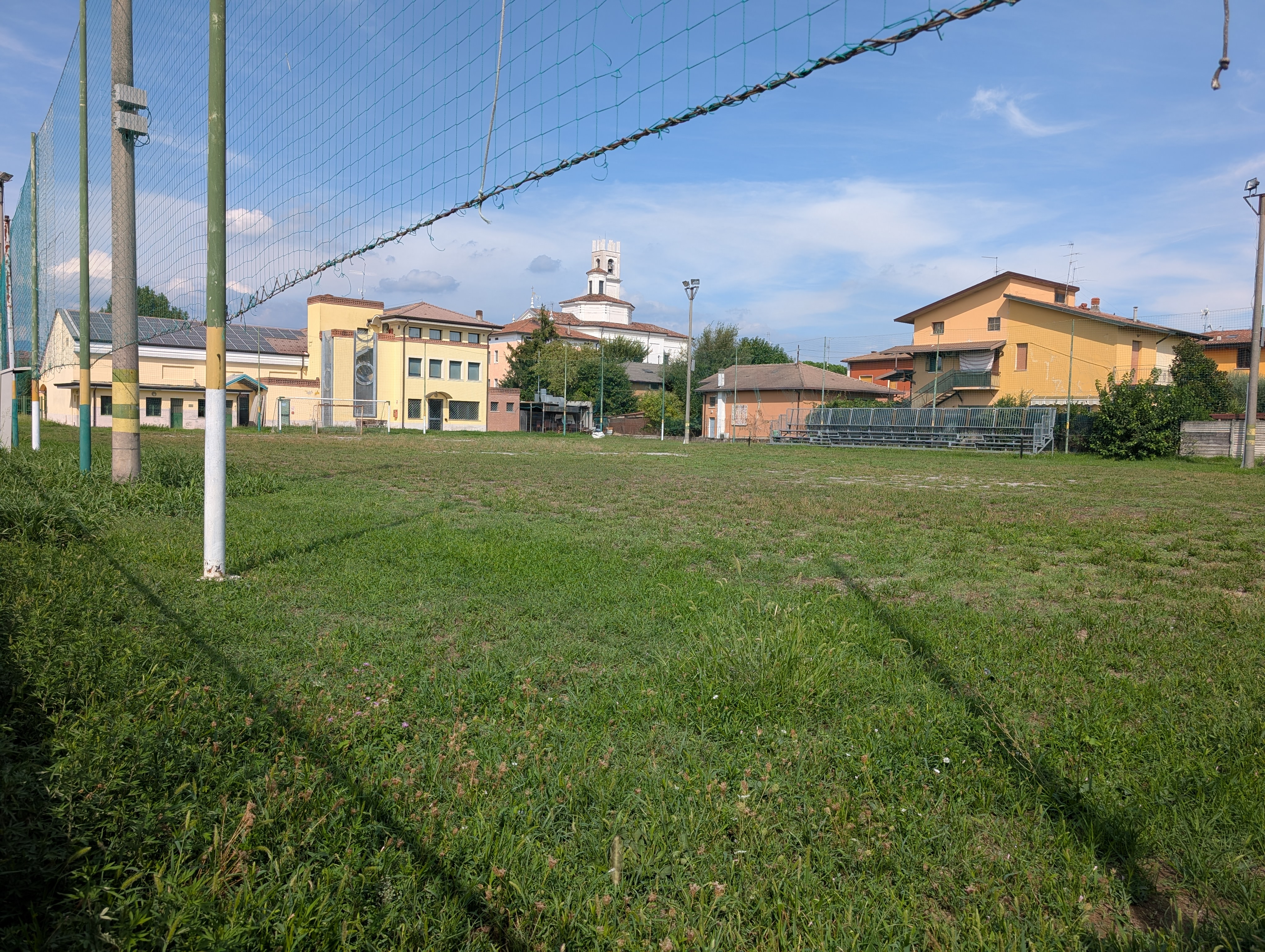 Una porta e la rete del campo sportivo con l’oratorio e il campanile sullo sfondo
