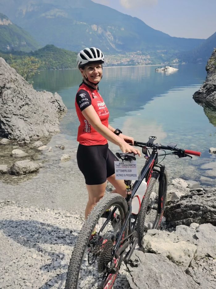 Simona Casana accanto alla sua mountain bike sulla riva di un lago di montagna, in maglia rossa e casco