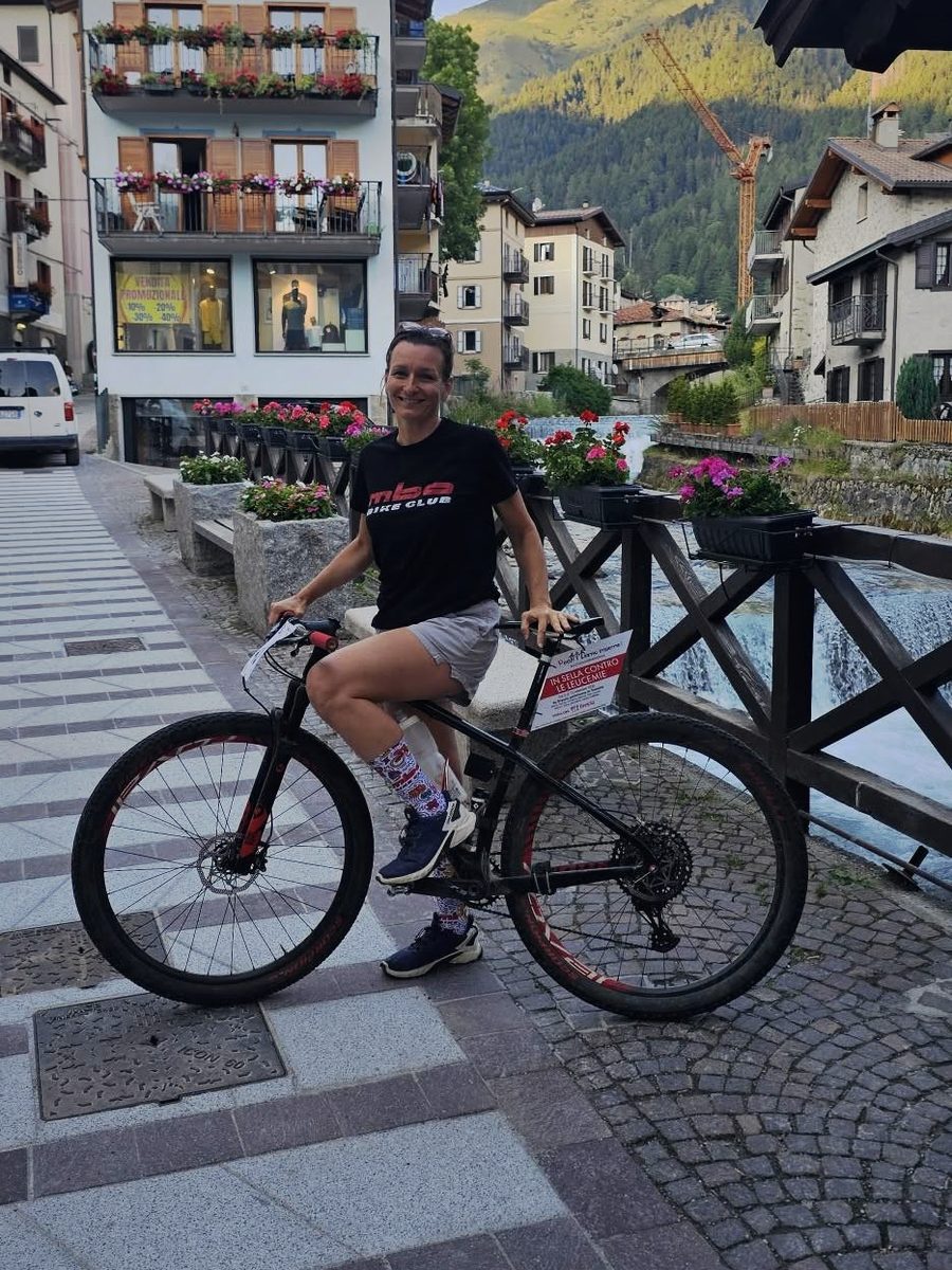 Simona Casana in sella alla bici in un paese di montagna, accanto a un torrente e a una casa con i balconi fioriti